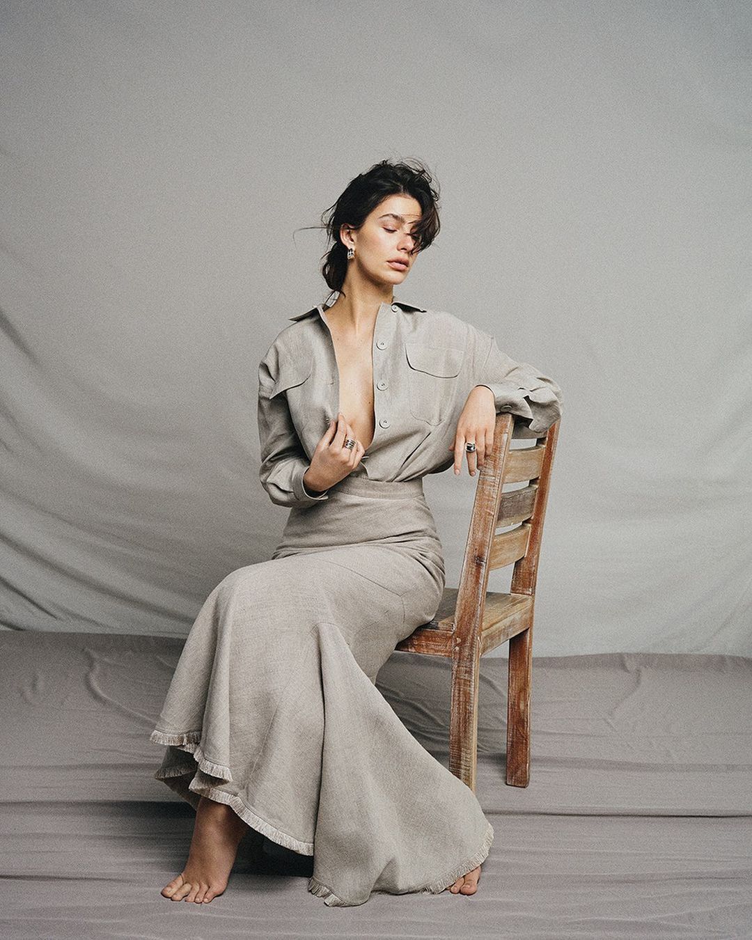 Brunette woman in neutral linen shirt and skirt sitting on a wood chair in front of a grey backdrop in a fashion photography studio.