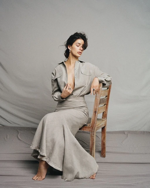 Brunette woman in neutral linen shirt and skirt sitting on a wood chair in front of a grey backdrop in a fashion photography studio.