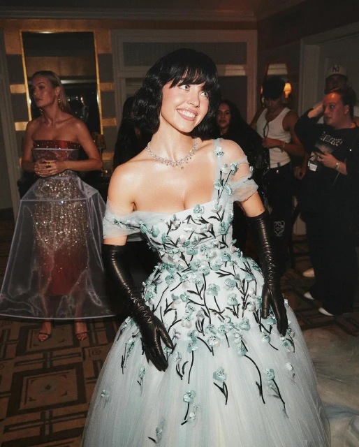 Woman smiles in a formal setting wearing a light blue tulle gown with floral embroidery, black opera gloves, and a statement necklace.