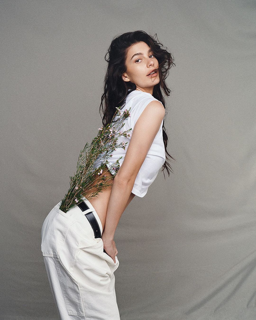 Young woman in white crop top and pants poses with small white flowers against a plain background in a studio shot.