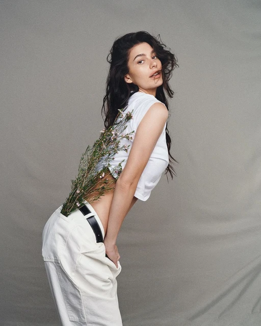 Young woman in white crop top and pants poses with small white flowers against a plain background in a studio shot.