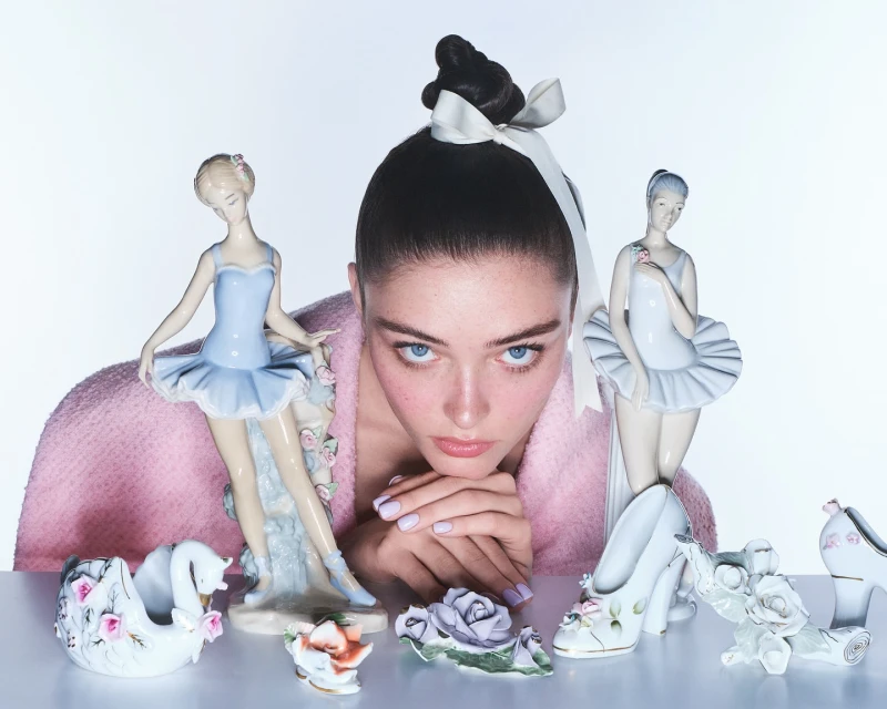 Portrait of a young woman with a high bun and ribbon, surrounded by decorative ballerina figurines and porcelain rose ornaments.