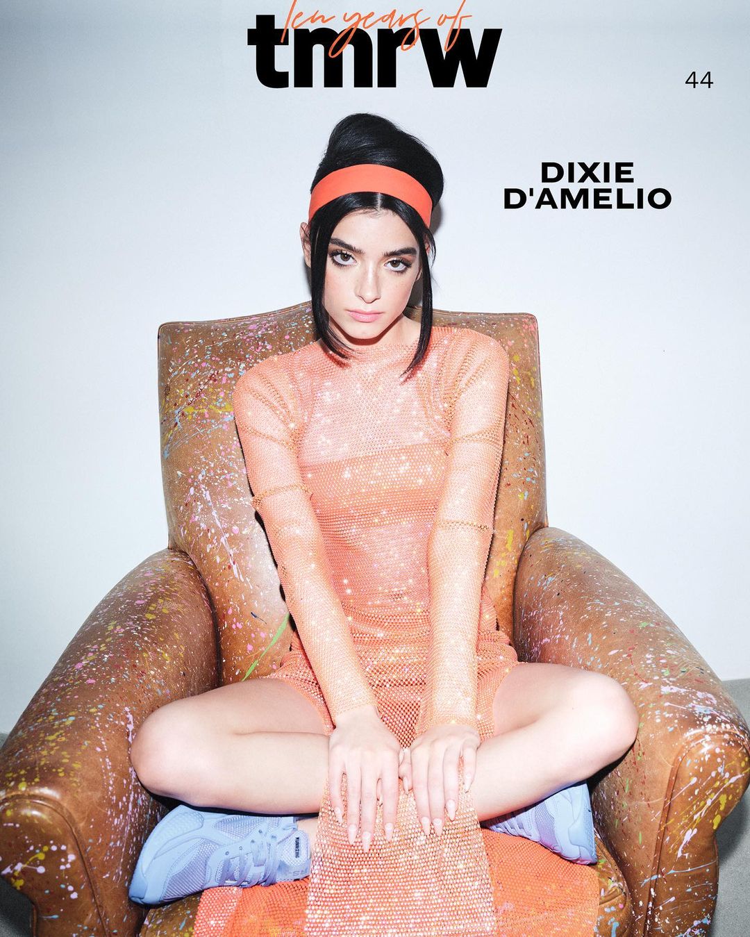 Dixie D'Amelio seated in an armchair wearing a sheer orange dress, orange headband, and blue sneakers against a plain background.