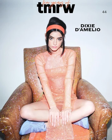 Dixie D'Amelio seated in an armchair wearing a sheer orange dress, orange headband, and blue sneakers against a plain background.