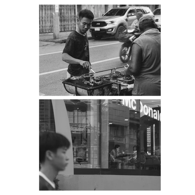 Two men work at outdoor grill on city street, viewed in black and white. McDonald's restaurant reflects in a nearby building window.