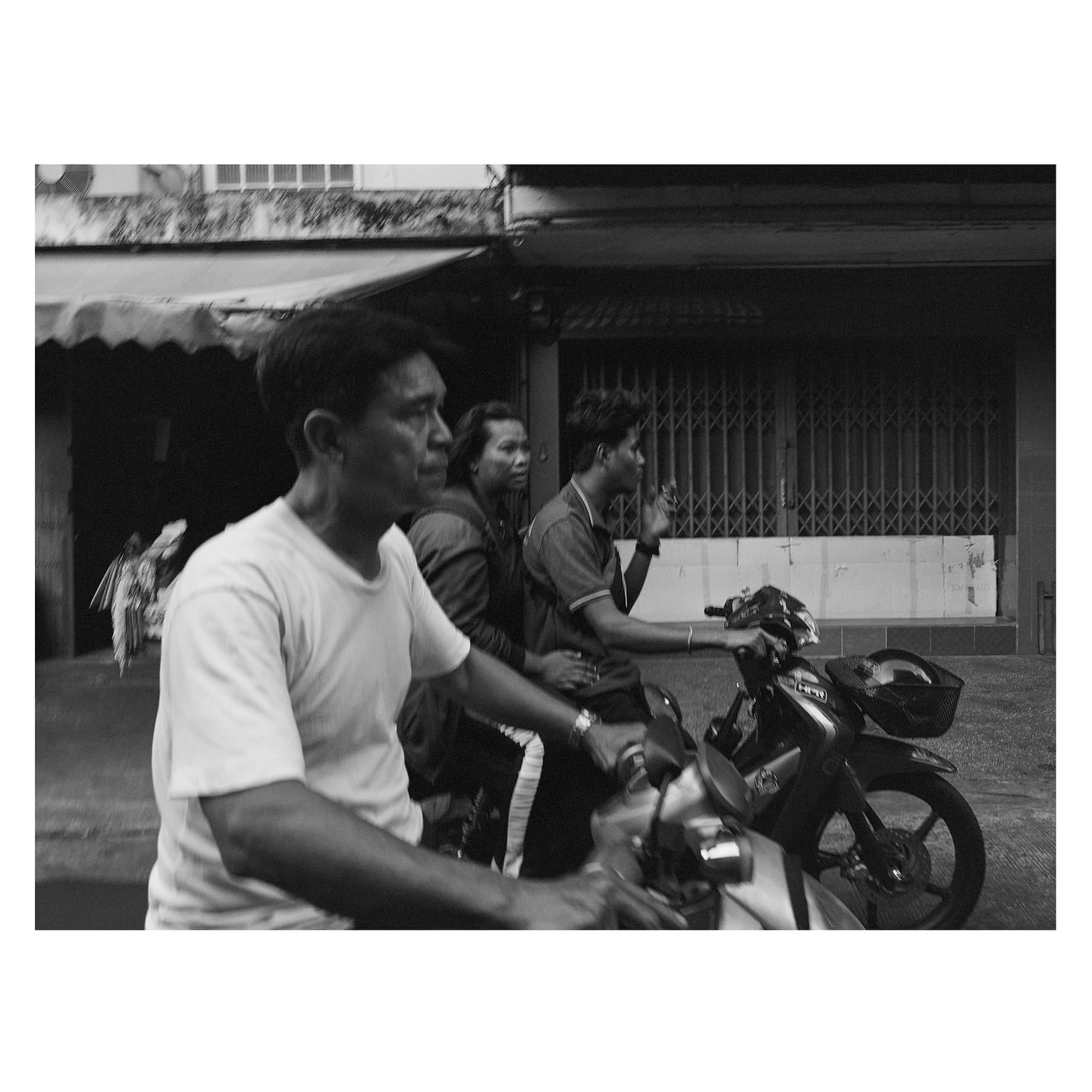 Three people ride a black motorcycle on a city street in a black and white photo. Buildings and awnings line the background.