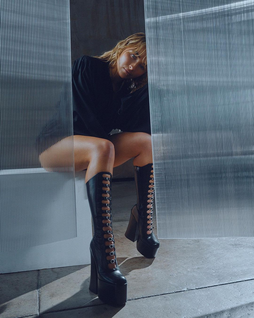 A woman is sitting in tall, black, platform boots with an edgy fashion style, against a textured background, looking at the camera.