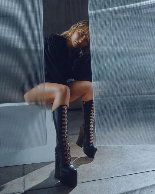 A woman is sitting in tall, black, platform boots with an edgy fashion style, against a textured background, looking at the camera.