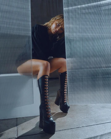 A woman is sitting in tall, black, platform boots with an edgy fashion style, against a textured background, looking at the camera.
