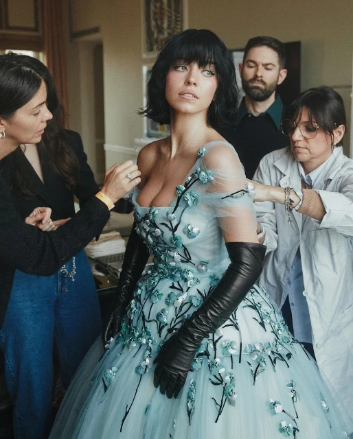Fashion shoot featuring a woman in a light blue, floral-applique dress being fitted by two stylists, with a man standing in the background.