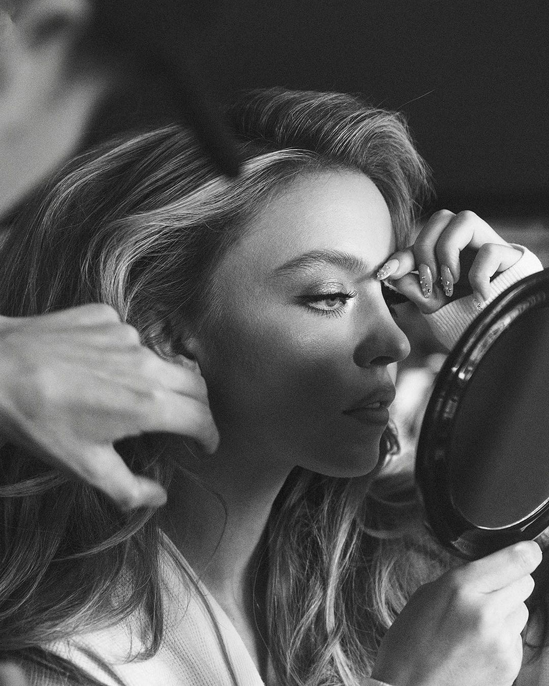 Black and white portrait of a woman getting her hair and makeup done. She's holding a small round mirror and looking into it.