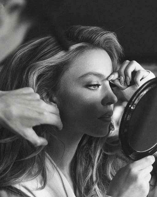 Black and white portrait of a woman getting her hair and makeup done. She's holding a small round mirror and looking into it.