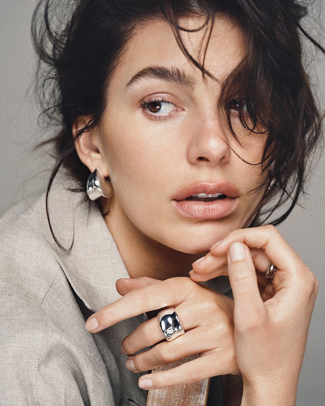 Close-up portrait of an adult woman with dark hair and neutral makeup wearing statement earrings and a ring, shot in soft studio lighting.