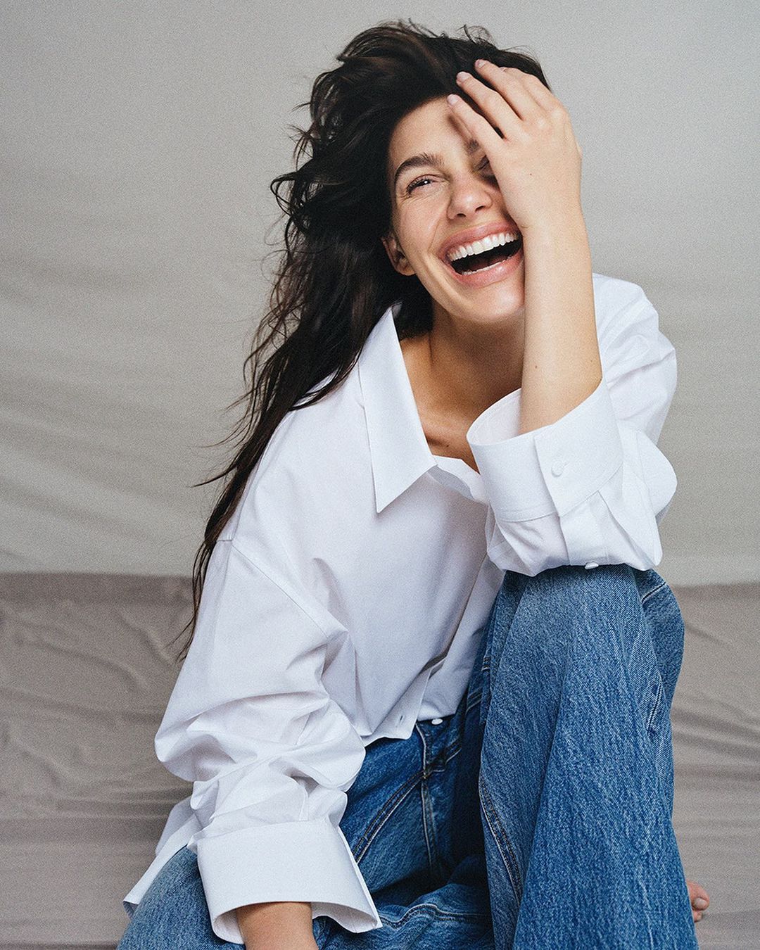 A woman with dark hair throws her head back and laughs, covering one eye, wearing a white collared shirt and blue jeans.