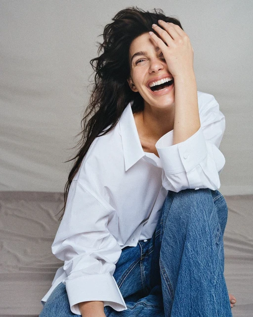A woman with dark hair throws her head back and laughs, covering one eye, wearing a white collared shirt and blue jeans.