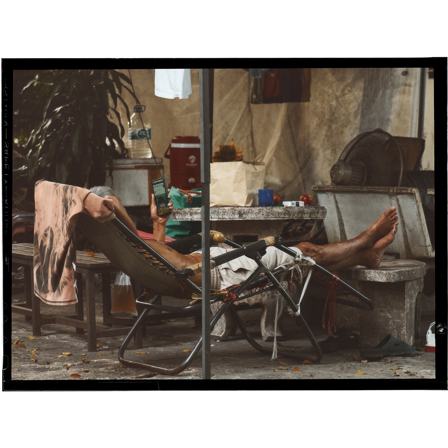An older person relaxing in a folding chair outside, using a cell phone. The scene is framed by outdoor furniture and a shaded backdrop.