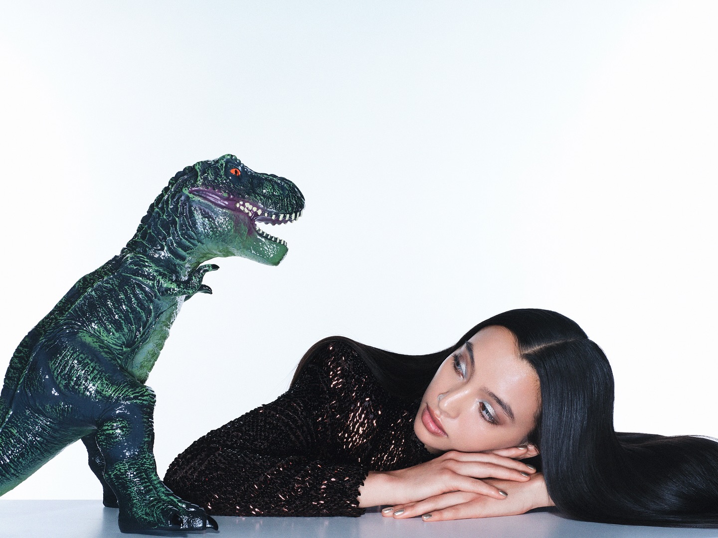 Woman in a sequined top lies next to a green dinosaur toy against a white backdrop, creating a surreal studio composition.