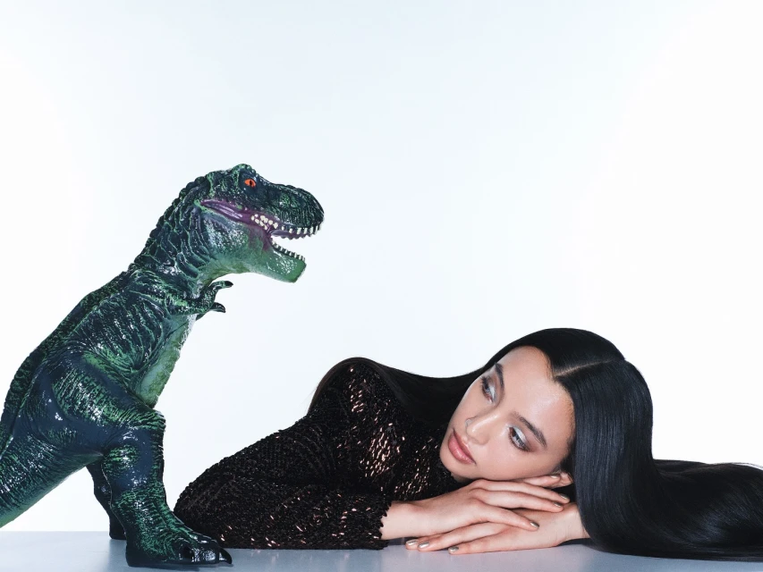Woman in a sequined top lies next to a green dinosaur toy against a white backdrop, creating a surreal studio composition.