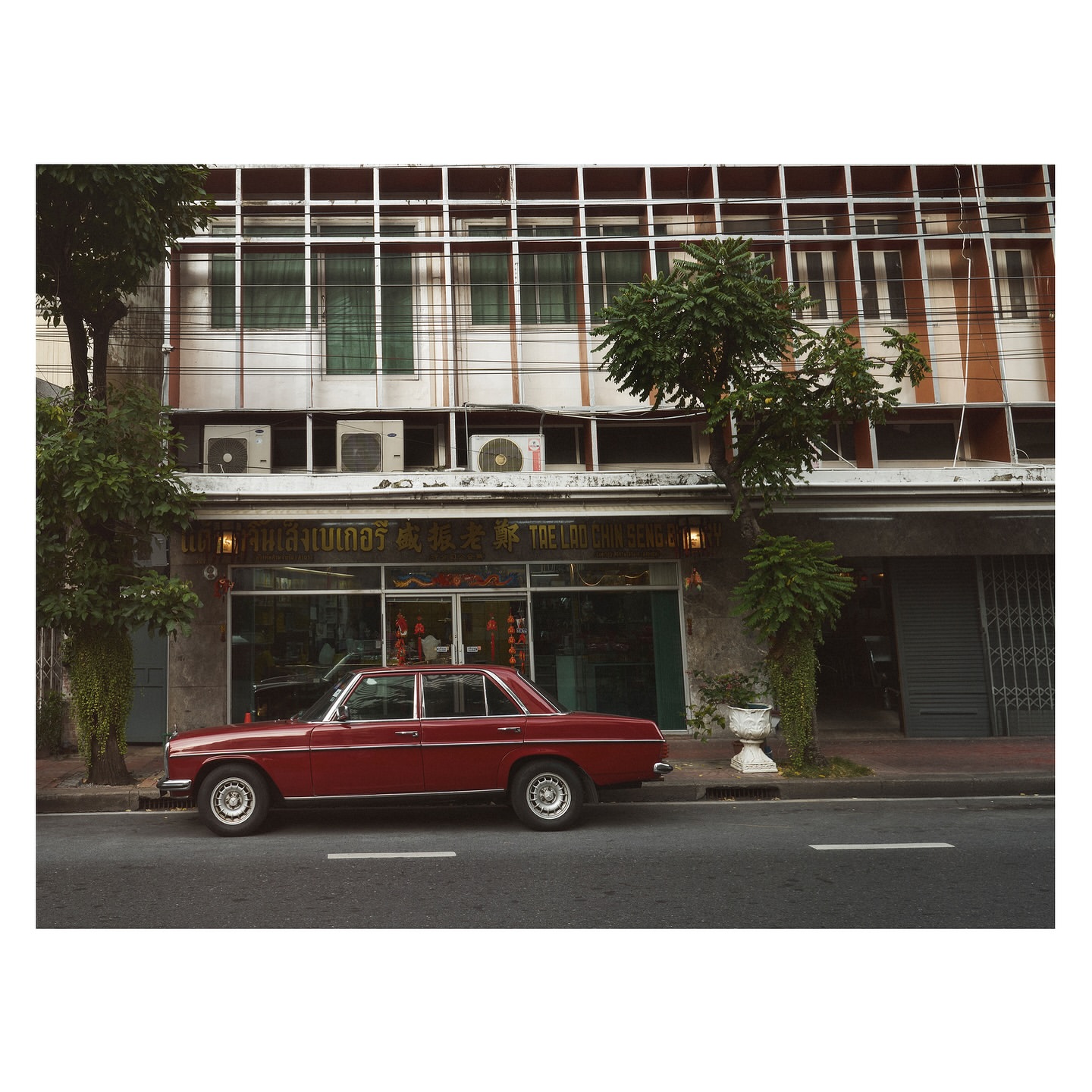 A vintage red car parked along a city street in front of an older commercial building with glass windows.