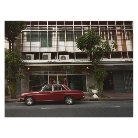 A vintage red car parked along a city street in front of an older commercial building with glass windows.