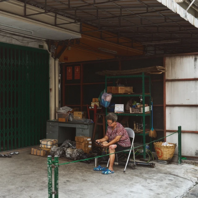 Elderly woman sitting on a chair outdoors on the sidewalk outside of a building with stacked boxes, storage, and a metal gate.