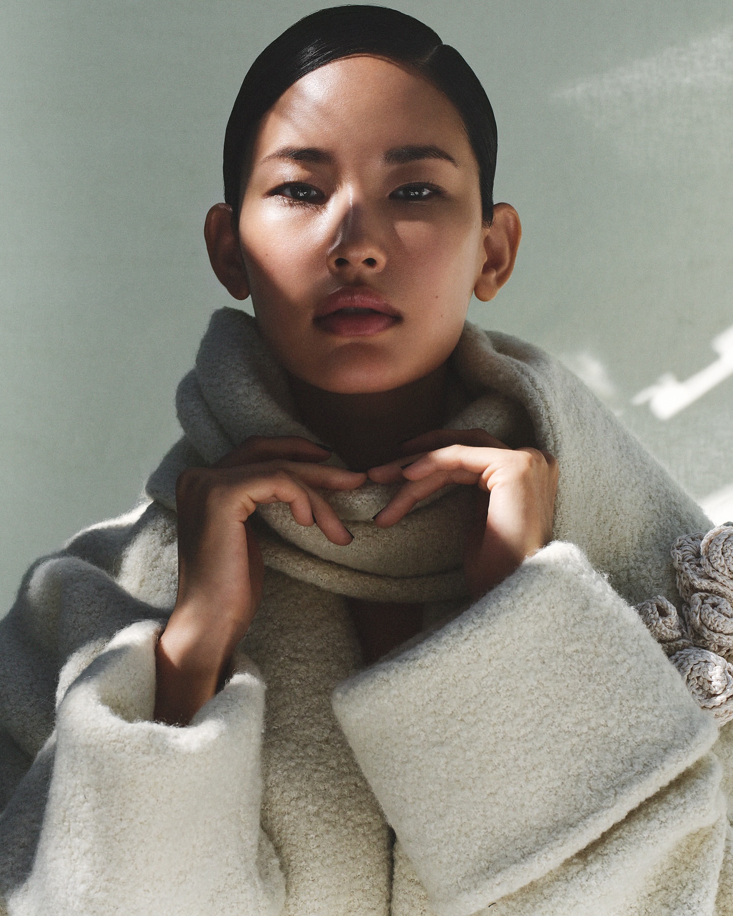 Portrait of an Asian woman wearing a white textured coat, hands near her neck, against a soft-colored background, fashion editorial.