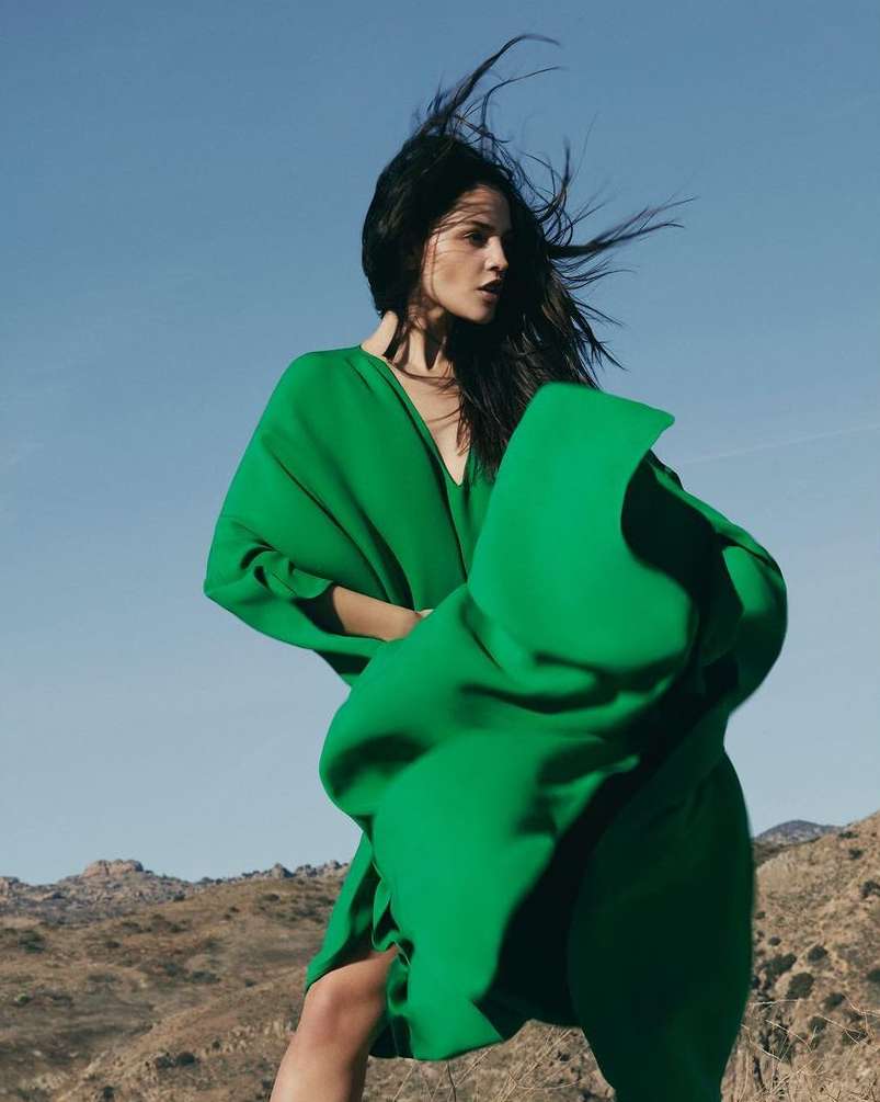 Woman with long dark hair wearing a vibrant green dress against a clear blue sky and arid landscape background