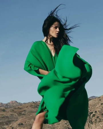Woman with long dark hair wearing a vibrant green dress against a clear blue sky and arid landscape background