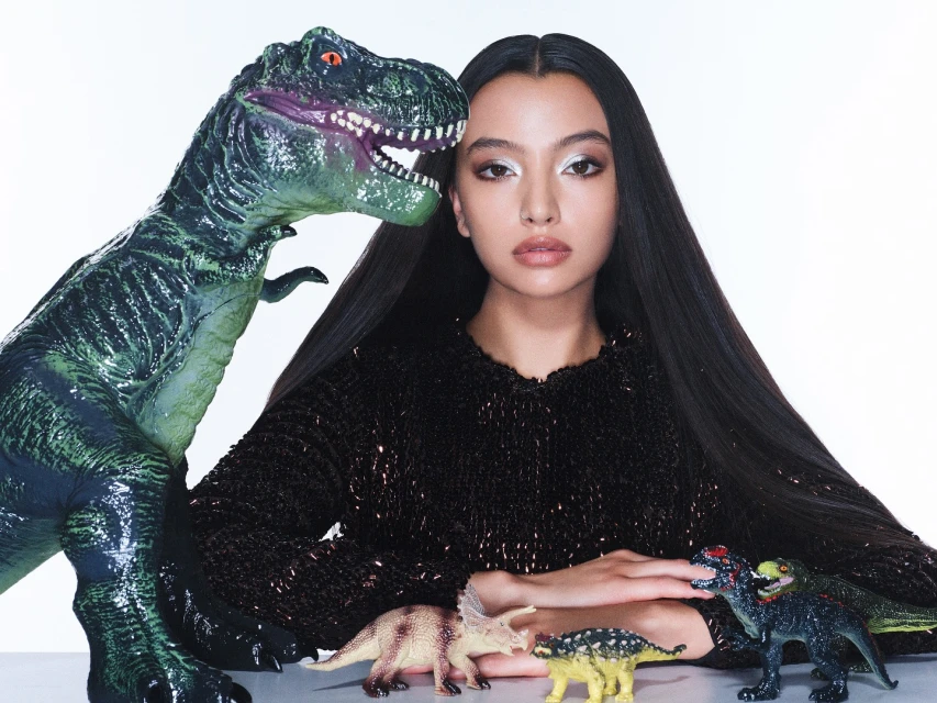 Elegant woman poses with toy dinosaurs in a bright studio setting, wearing a dark, shimmering top and silver eyeshadow. 