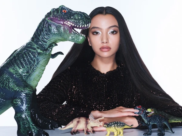 Elegant woman poses with toy dinosaurs in a bright studio setting, wearing a dark, shimmering top and silver eyeshadow. 