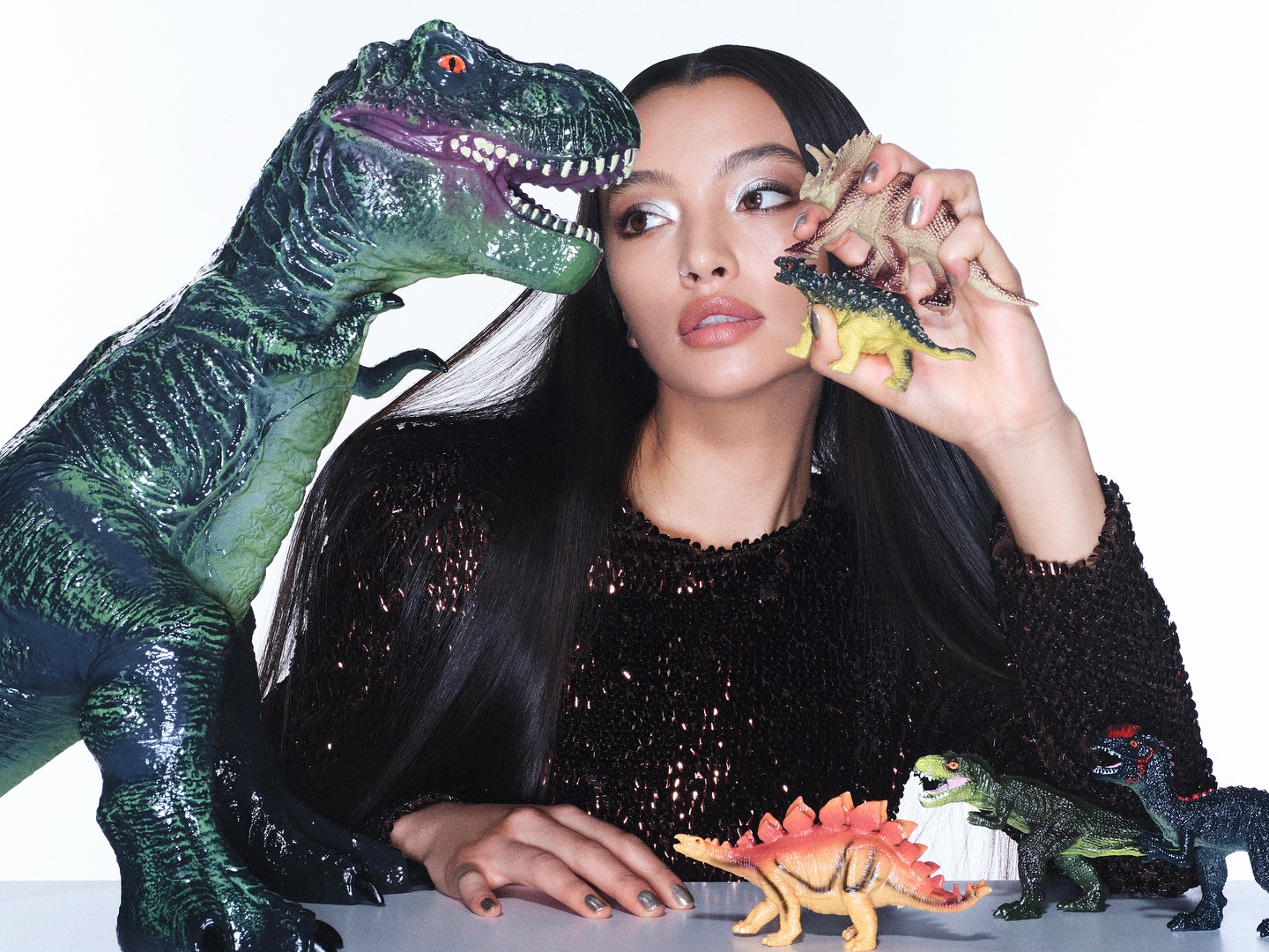 Attractive Asian woman holds miniature dinosaurs in a studio environment, standing next to a large toy T-Rex. Fashion and beauty shot.