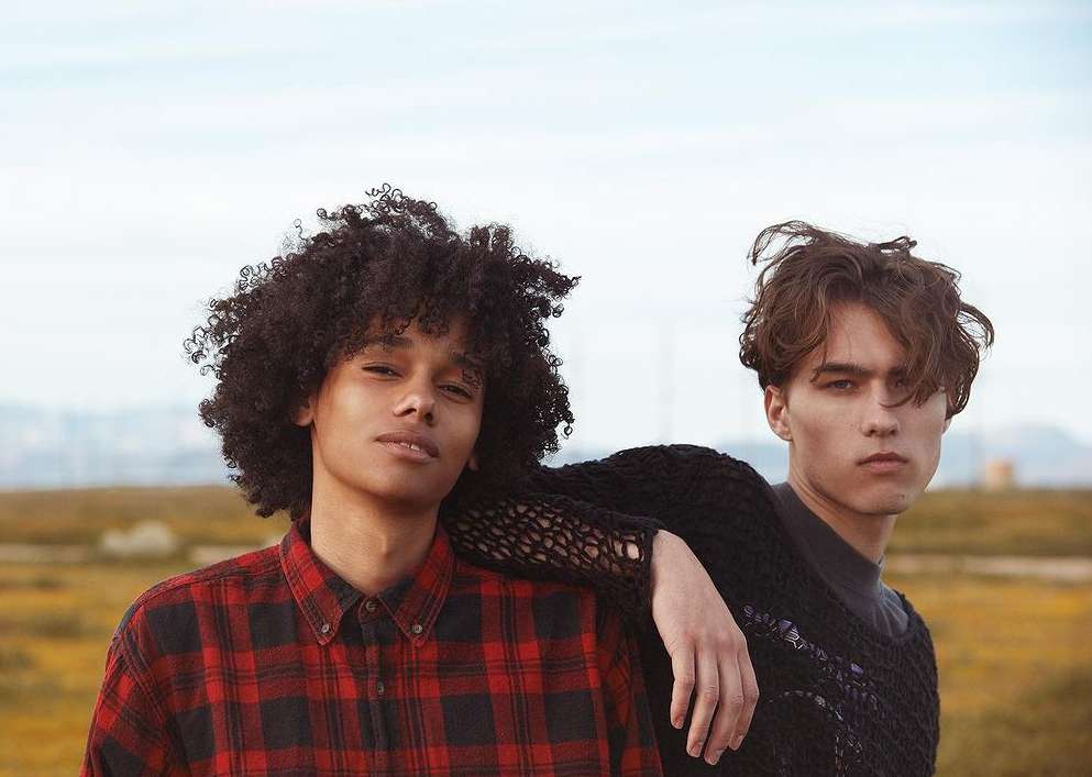 Two young adult fashion models pose outdoors in knit sweaters and a red plaid shirt against a background of grassy fields and sky.