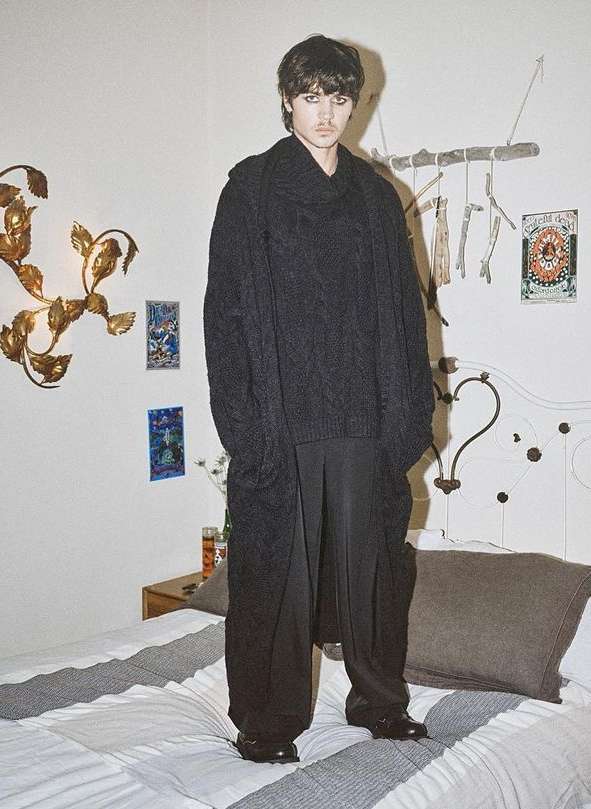 Young adult stands on bed in oversized black knit sweater, wide-leg pants, and shiny shoes in bright indoor fashion shot.