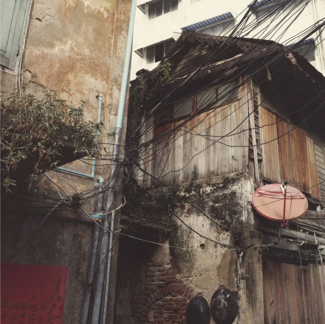 Exterior of an old weathered wooden building in Bangkok, Thailand, with power lines, brick walls, and plants in an urban alleyway.