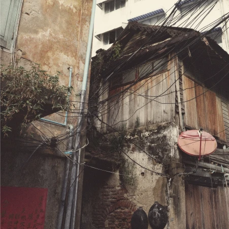 Exterior of an old weathered wooden building in Bangkok, Thailand, with power lines, brick walls, and plants in an urban alleyway.