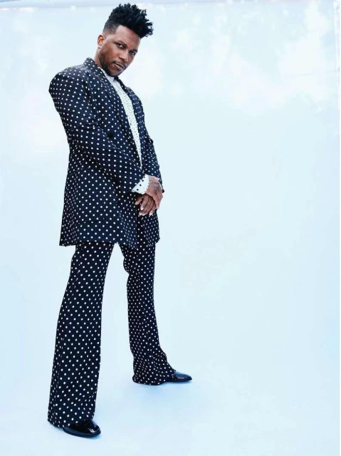 An adult male stands in a full-length shot, wearing a black and white polka dot suit against a light blue seamless background.