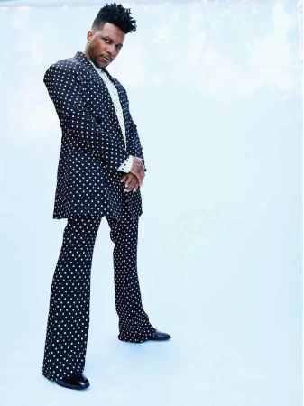 An adult male stands in a full-length shot, wearing a black and white polka dot suit against a light blue seamless background.