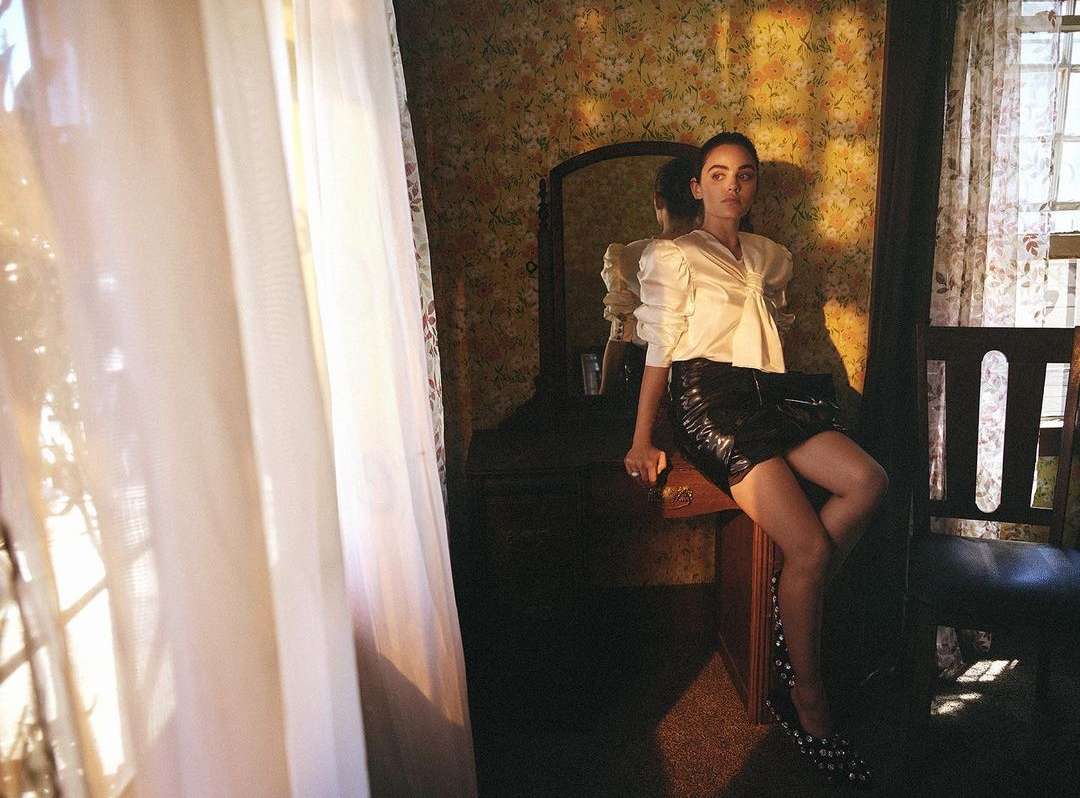 Woman in a white satin top and black mini skirt poses inside a room with floral wallpaper near a window with sheer curtains.