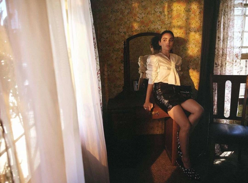 Woman in a white satin top and black mini skirt poses inside a room with floral wallpaper near a window with sheer curtains.