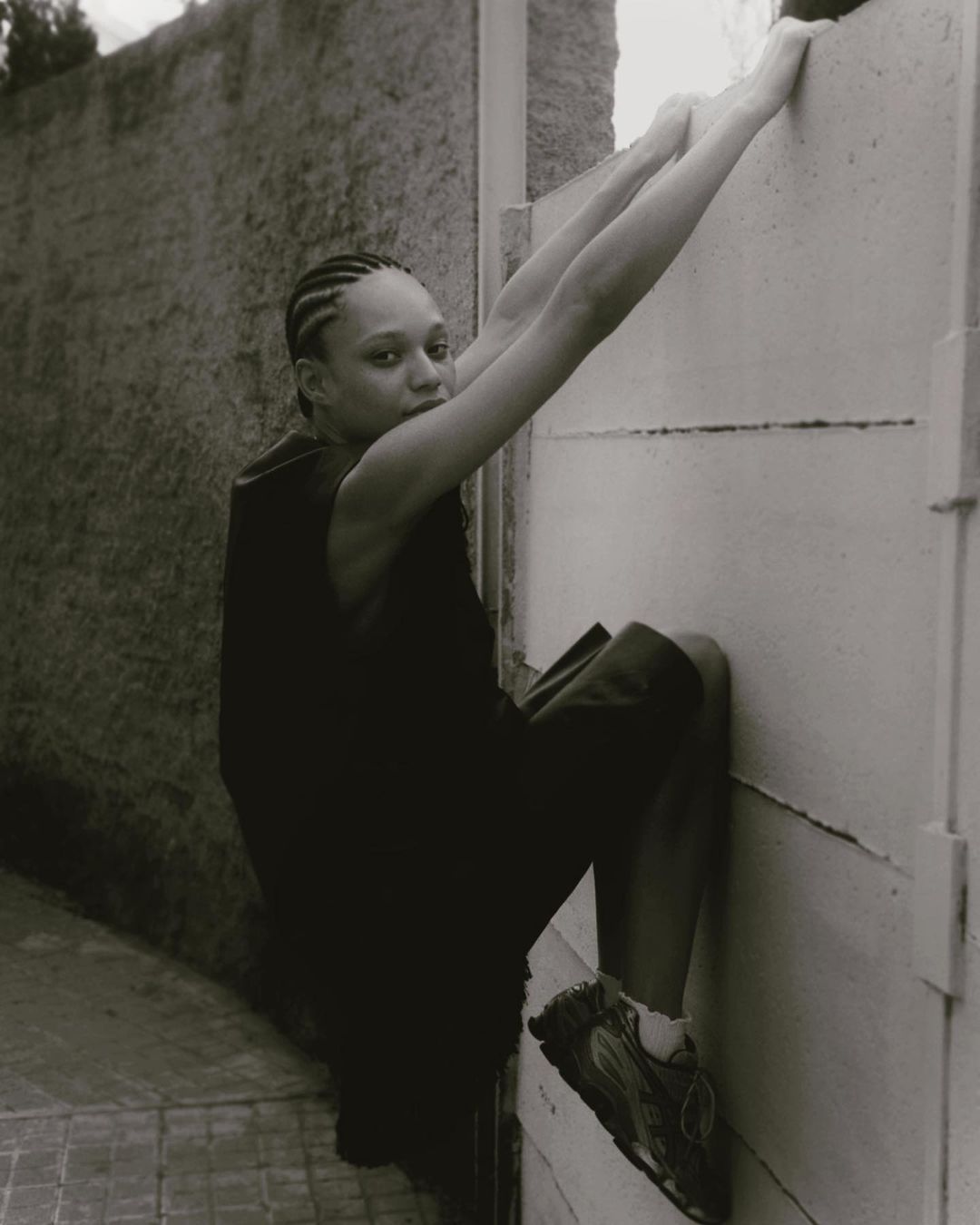Black and white portrait of an adult female climbing a wall outdoors, wearing a tank top and shorts, with braided hair and athletic shoes.