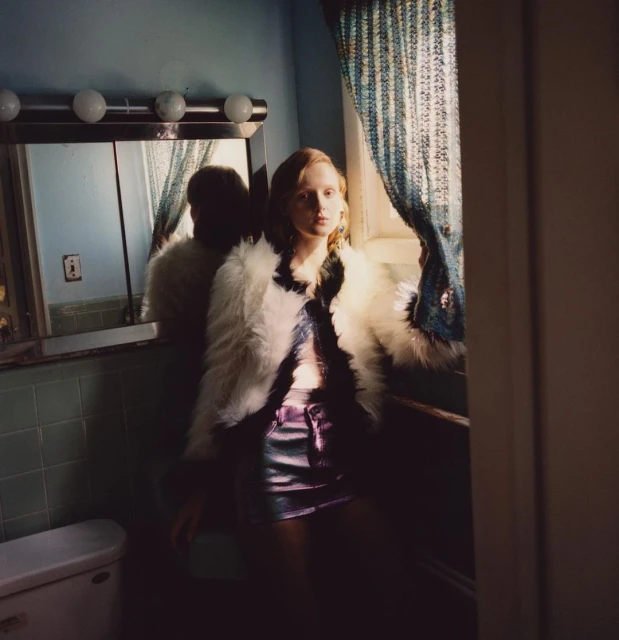 Young woman wearing a fur coat and shorts is posed indoors near a window with blue curtains and a mirror in the background.