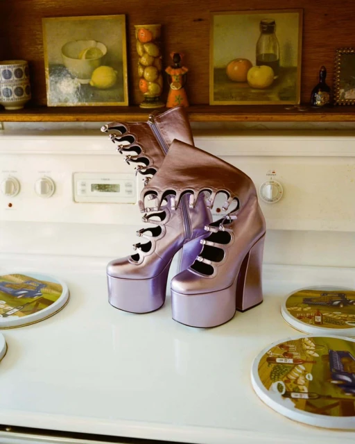 Platform boots in lilac metallic on a white stove top with vintage artwork visible on the shelf above, indoor kitchen setting.