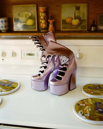 Platform boots in lilac metallic on a white stove top with vintage artwork visible on the shelf above, indoor kitchen setting.
