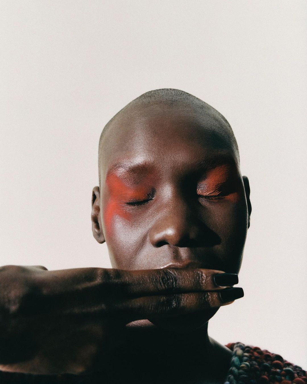 Close-up portrait of a bald Black model with red eye makeup holding fingers across lips by Dennis Stenild