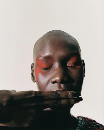 Close-up portrait of a bald Black model with red eye makeup holding fingers across lips by Dennis Stenild