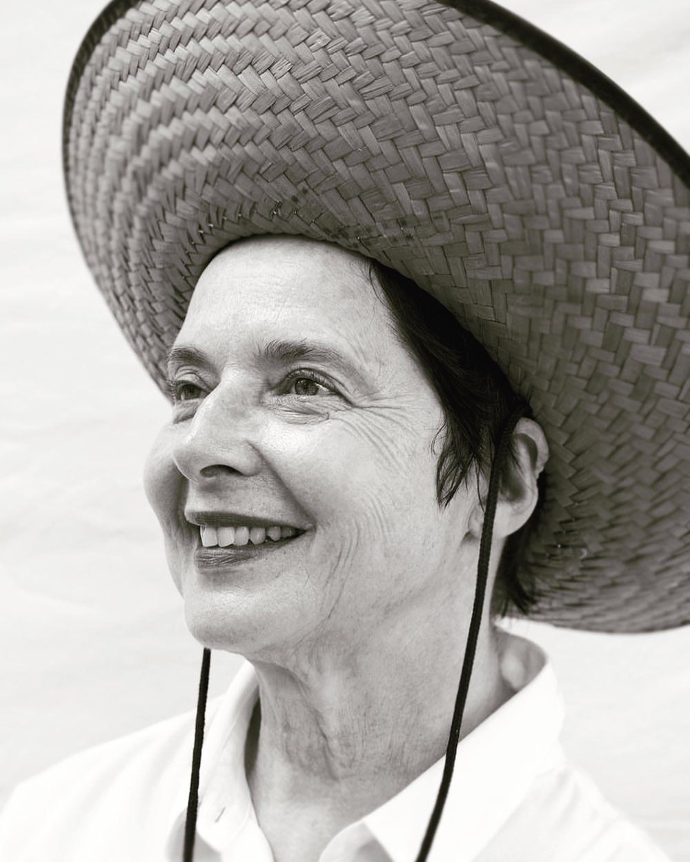Monochrome portrait of a smiling, mature woman wearing a straw hat, looking up and to the left, with a light, solid background.