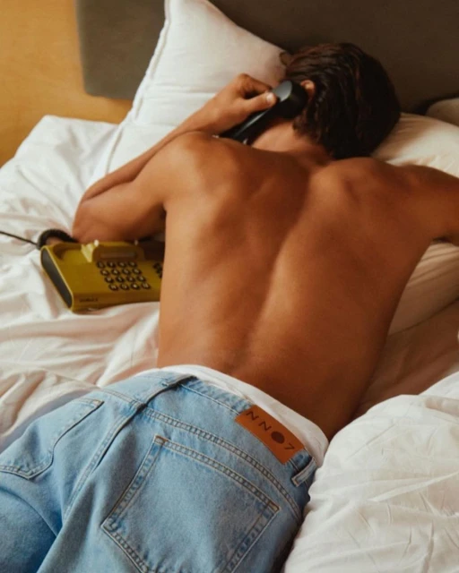 Shirtless man lying on bed talking on rotary phone in bedroom setting. Light blue jeans and white sheets visible.