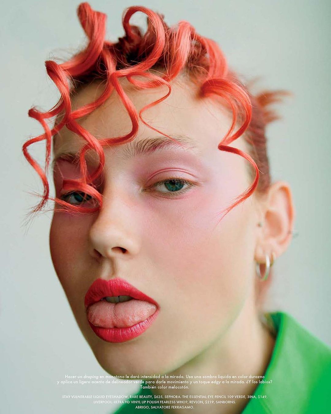Close-up of a young fair-skinned woman with coral curly hair and bright makeup, sticking her tongue out, wearing a green jacket.