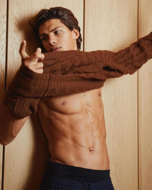 Athletic man with brown hair poses shirtless with a brown cable knit sweater draped on his shoulder, against a light wood panel wall.