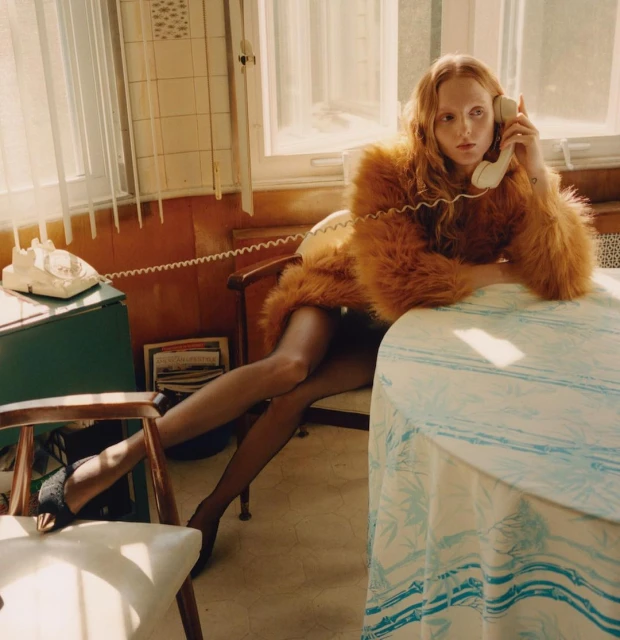A woman in an orange faux fur coat sits at a table talking on a vintage telephone in an indoor setting near a window.